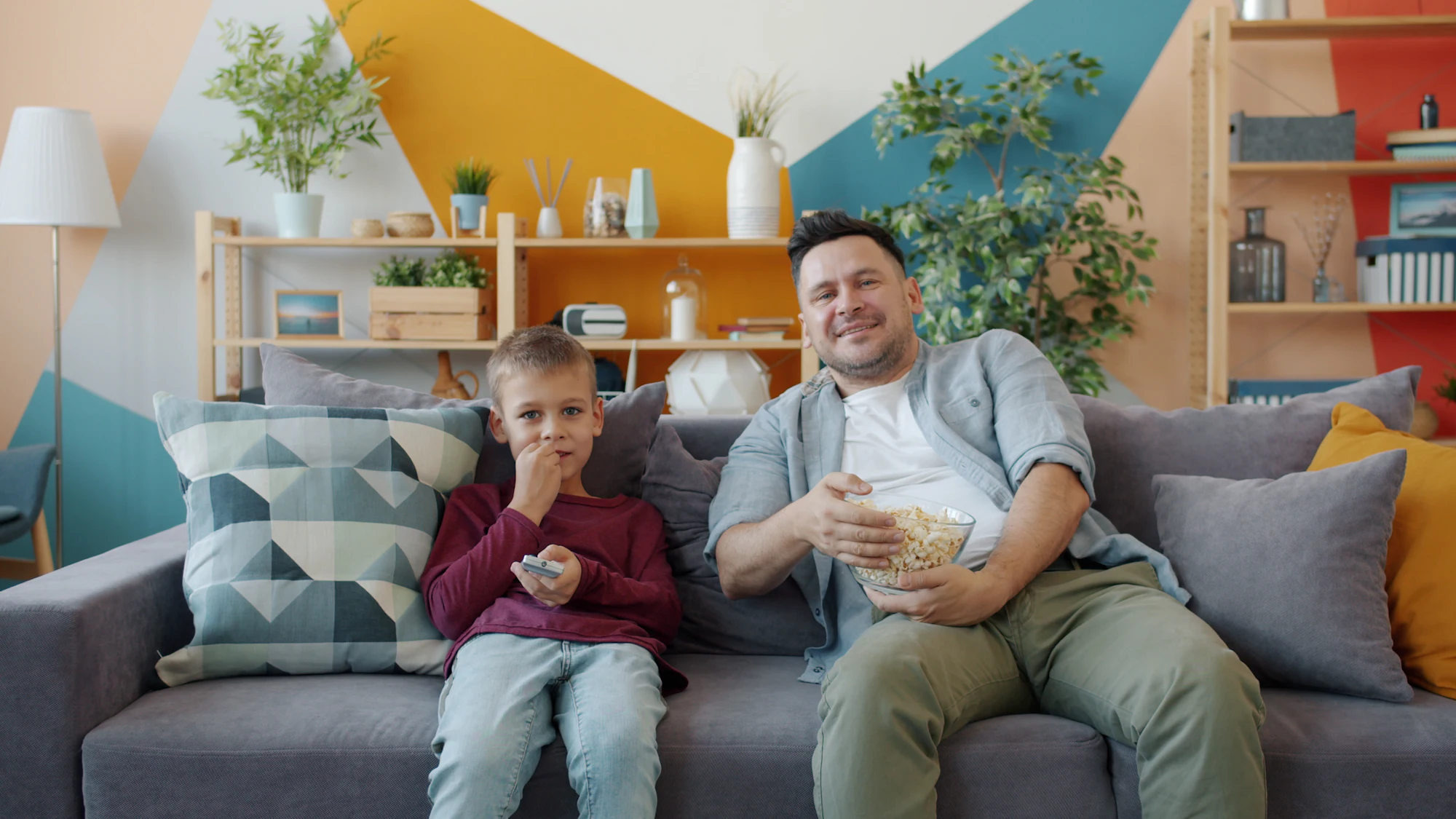 Father and son relaxing on a sofa with popcorn while watching TV at home